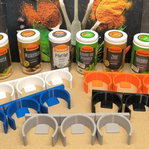 May include: A set of eight spice jars with various labels, including Curry, Muskatnuss, Pfeffer, Krauter der Provence, Zimt, and Oregano. The jars are arranged in two rows of four, with a white background. The jars are all the same size and shape, and they have a clear glass body with a black lid. The labels are all white with black text.