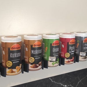 May include: A row of eight spice jars with white lids and black labels. The labels have the brand name "Ostmann" and the name of the spice in German. The jars are mounted on a wall-mounted rack.