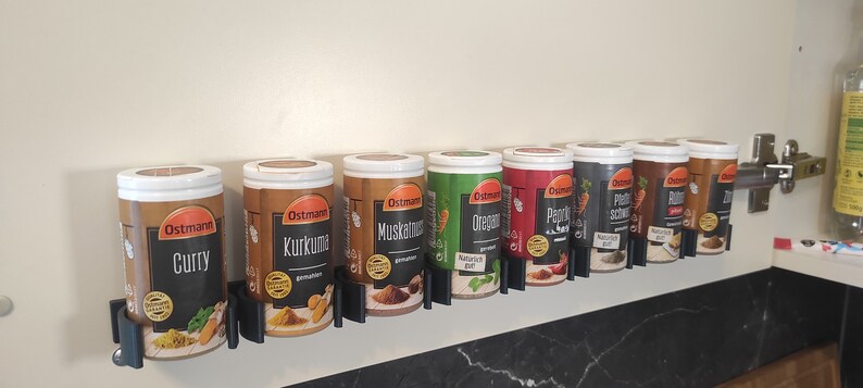 May include: A row of eight spice jars with white lids and black labels. The labels have the brand name "Ostmann" and the name of the spice in German. The jars are mounted on a wall-mounted rack.