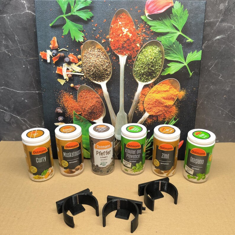 May include: Seven small, round, white spice jars with black lids and green and white labels. The labels have the brand name "Ostmann" and the name of the spice in German. The jars are arranged in a row on a brown surface. The background is a close-up of a canvas with spoons filled with various spices.