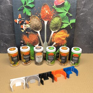 May include: Six spice jars with labels in German, including Curry, Muskatnuss, Pfeffer,  Kr&auml;uter der Provence, Zimt, and Oregano. The jars are arranged in a row on a brown surface.  Below the jars are six spice rack holders in white, gray, black, orange, and blue.