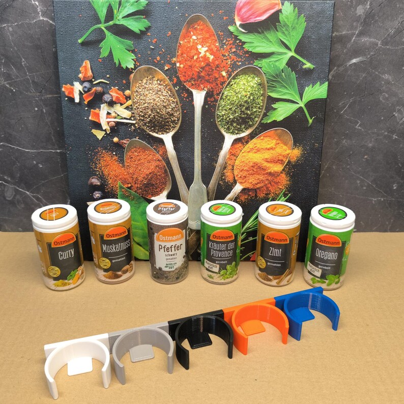 May include: Six spice jars with labels in German, including Curry, Muskatnuss, Pfeffer,  Kr&auml;uter der Provence, Zimt, and Oregano. The jars are arranged in a row on a brown surface.  Below the jars are six spice rack holders in white, gray, black, orange, and blue.