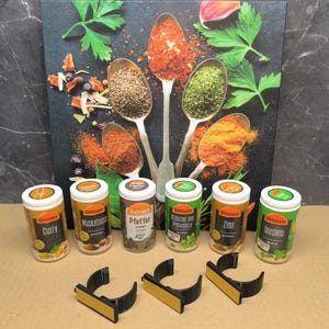May include: Six small spice jars with black lids and orange labels. The labels have the brand name "Ostmann" and the spice name, including Curry, Muskatnuss, Pfeffer, Krauter der Provence, Zimt, and Oregano. The jars are arranged in a row on a wooden surface.