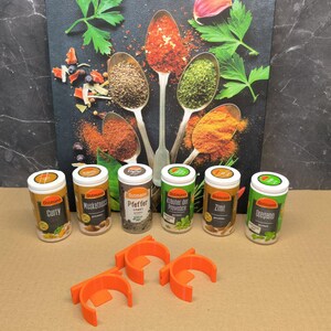 May include: Six small round spice jars with orange lids and labels. The labels have the brand name "Ostmann" and the spice name, such as "Curry", "Muskatnuss", "Pfeffer", "Kr&auml;uter der Provence", "Zimt", and "Oregano". The jars are arranged in a row on a white surface. There are two orange plastic spice rack holders in the foreground.