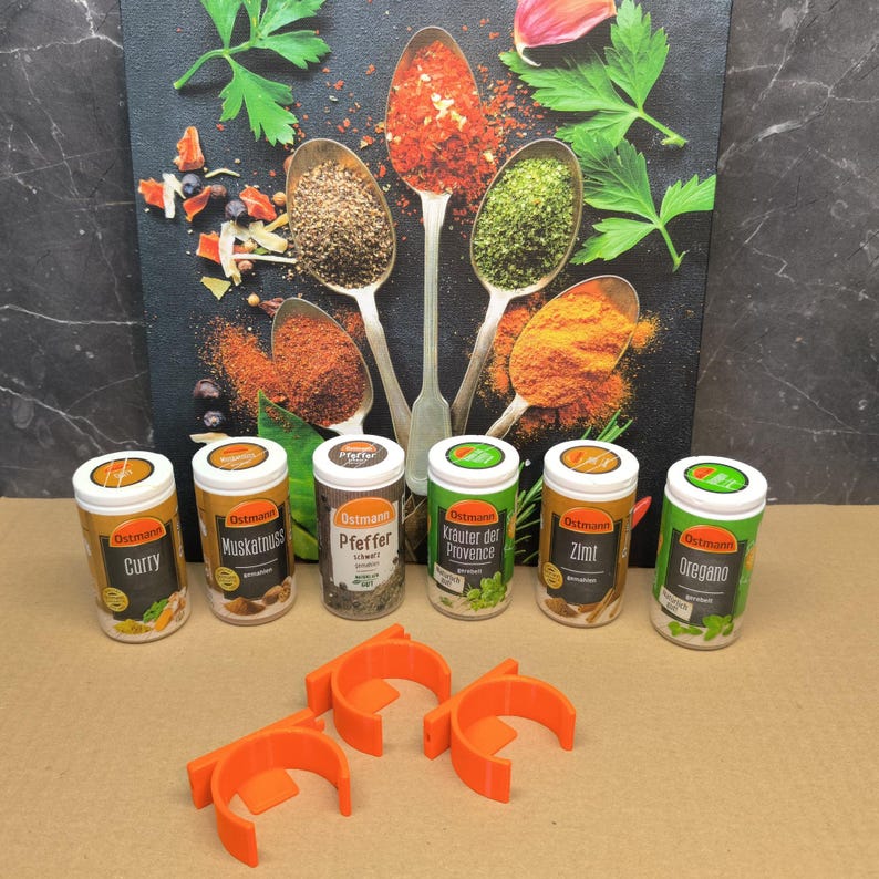 May include: Six small round spice jars with orange lids and labels. The labels have the brand name "Ostmann" and the spice name, such as "Curry", "Muskatnuss", "Pfeffer", "Kr&auml;uter der Provence", "Zimt", and "Oregano". The jars are arranged in a row on a white surface. There are two orange plastic spice rack holders in the foreground.