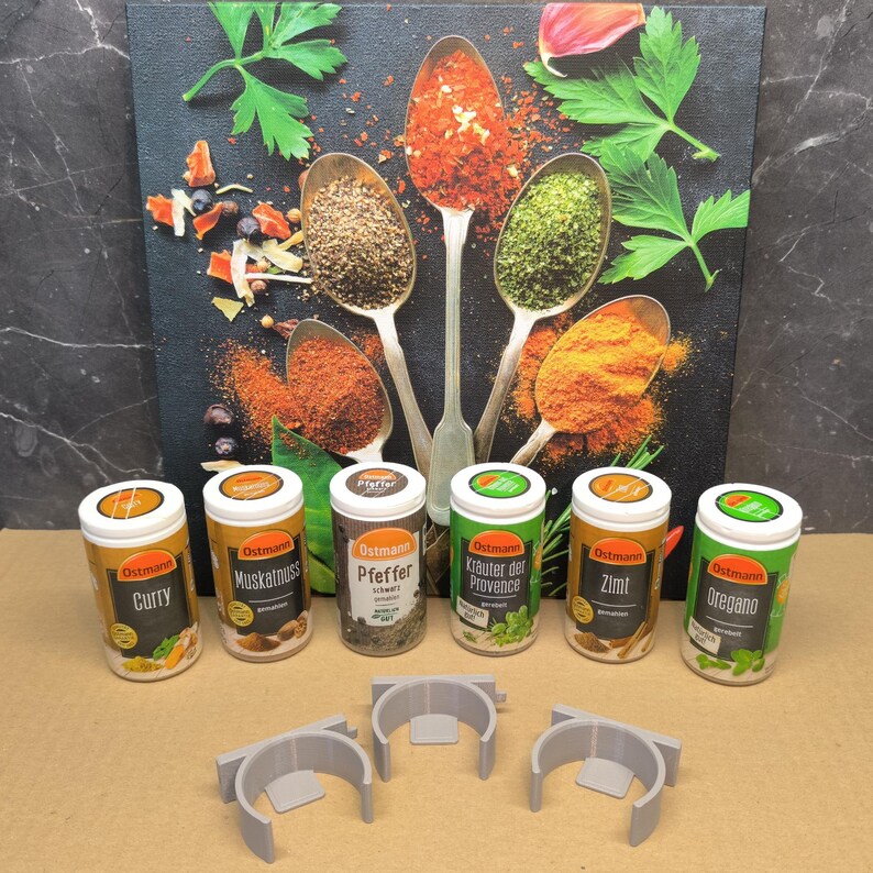May include: Seven spice jars with white lids and green labels. The labels have the brand name "Ostmann" and the name of the spice in German. The spices are curry, nutmeg, pepper, herbs of Provence, cinnamon, and oregano. The jars are sitting on a light brown surface in front of a canvas with a picture of spoons filled with spices.