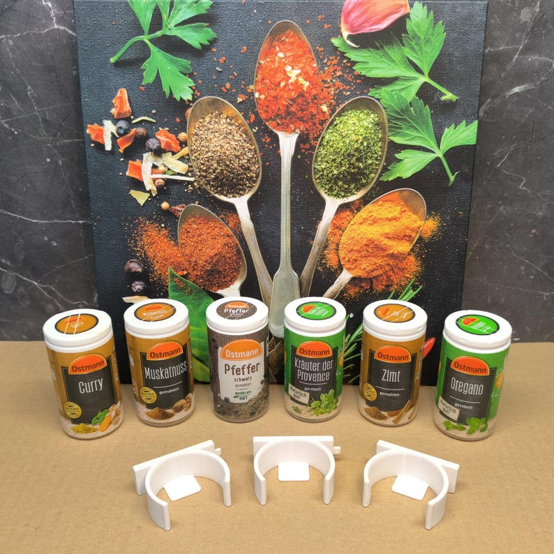 May include: Seven spice jars with white lids and orange labels. The labels have the brand name "Ostmann" and the name of the spice in black text. The spices are Curry, Muskatnuss, Pfeffer, Kr&auml;uter der Provence, Zimt, and Oregano. The jars are sitting on a white surface with six white plastic clips.