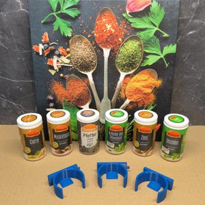 May include: Six small spice jars with various labels, including "Curry", "Muskatnuss", "Pfeffer", "Krauter der Provence", "Zimt", and "Oregano". The jars are arranged in a row on a brown surface. Three blue plastic spice rack holders are also visible in the image.