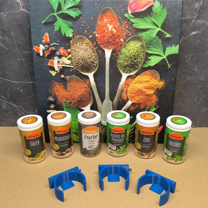 May include: Six small spice jars with various labels, including "Curry", "Muskatnuss", "Pfeffer", "Krauter der Provence", "Zimt", and "Oregano". The jars are arranged in a row on a brown surface. Three blue plastic spice rack holders are also visible in the image.