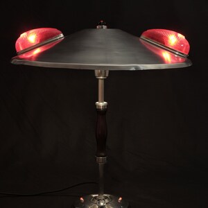 Può includere: Una lampada da tavolo argentata con un paralume a forma di piattino e tre luci rosse. La lampada ha una base in legno scuro e uno stelo argentato.