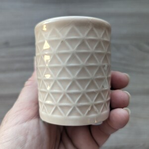May include: A white ceramic candle holder with a geometric pattern of triangles. The holder is cylindrical in shape and has a smooth, glossy finish.