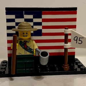May include: A Lego figurine of a scout wearing a tan hat and uniform, holding a brown staff and a silver cup. Behind the figure is a Lego American flag and a white flag with the number 95.