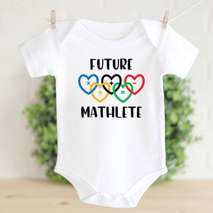 May include: White baby onesie with the text "FUTURE MATHLETE" in black. The text is surrounded by five interlocking hearts in the colors of the Olympic rings: blue, yellow, black, green, and red. Each heart has a different mathematical symbol: multiplication, addition, division, equals, and subtraction.