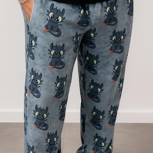 K&ouml;nnte beinhalten: Hellblaue Schlafanzughose mit einem sich wiederholenden Muster eines Cartoon-Drachencharakters. Der Drache ist schwarz mit gelben Augen und roten Akzenten. Die Hose hat Taschen und eine bequeme Passform.