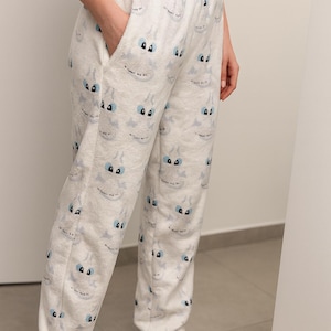 K&ouml;nnte beinhalten: Wei&szlig;e Schlafanzughose mit einem sich wiederholenden Cartoon-Monstergesichtsmuster in Hellblau und Schwarz. Die Hose hat einen Kordelzug in der Taille und Taschen. Die Person tr&auml;gt wei&szlig;e Socken.