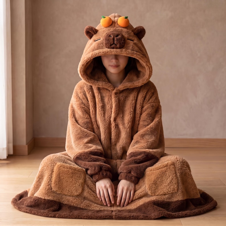 May include: A brown plush hooded onesie featuring a capybara face on the hood with two small orange fruit accents on top. The garment is made of soft, fuzzy material with long sleeves, a front opening, and two large square pockets on the front.