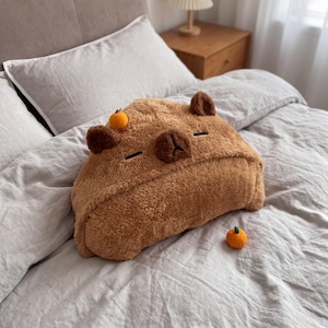 May include: A brown, plush capybara-shaped cushion with a soft, fuzzy texture. The cushion features dark brown ears, closed eyes, and a small nose. A tiny, bright orange fruit is attached to the top of the head, and a matching orange sits nearby.