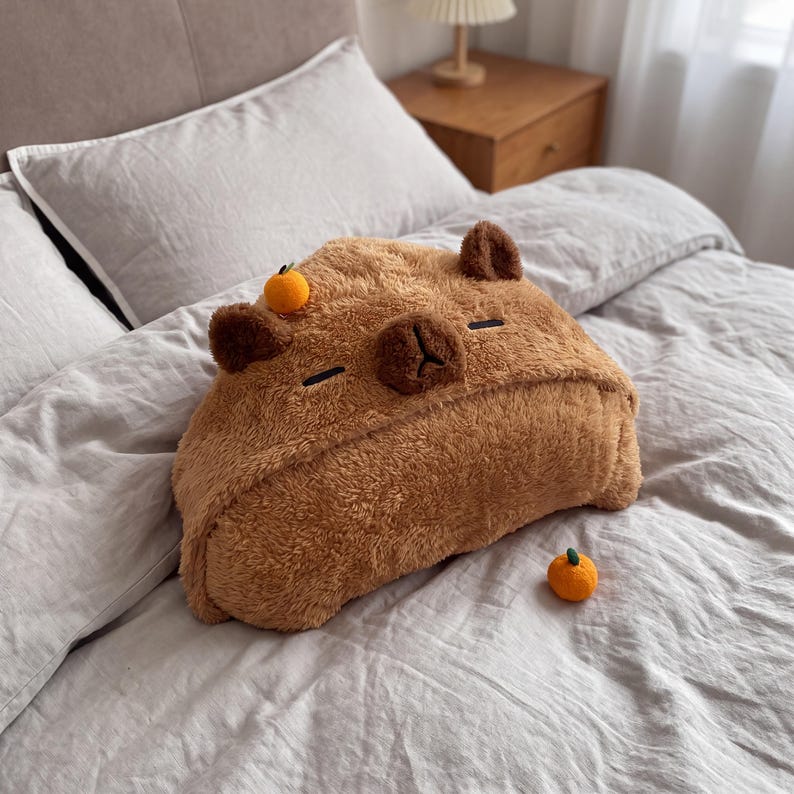 May include: A brown, plush capybara-shaped cushion with a soft, fuzzy texture. The cushion features dark brown ears, closed eyes, and a small nose. A tiny, bright orange fruit is attached to the top of the head, and a matching orange sits nearby.