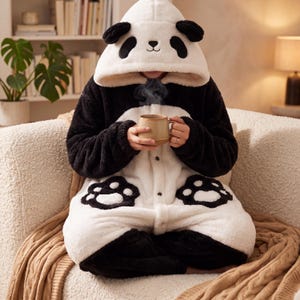 May include: A plush panda-themed onesie featuring a hood with black ears and panda facial features, a white torso with black sleeves, and black paw print appliqués on the front. The garment is made of soft, fuzzy material and includes buttons.
