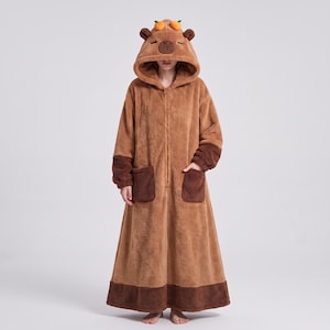 May include: A brown, full-length, plush hooded robe featuring a capybara face on the hood with two small orange accents on top. The garment includes a front zip closure, long sleeves with dark brown cuffs, and two large front patch pockets.