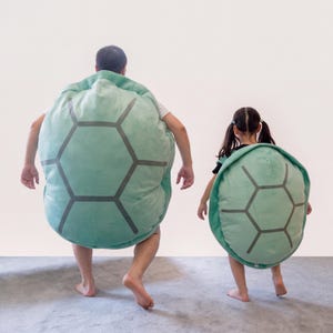 Op de afbeelding: Twee grote, ronde, mintgroene pluche schildpadschilden met donkergrijze zeshoekige patronen. Deze draagbare, zacht aanvoelende schilden zijn ontworpen om op de rug te dragen en hebben een afgeronde vorm die lijkt op een schildpadschild.