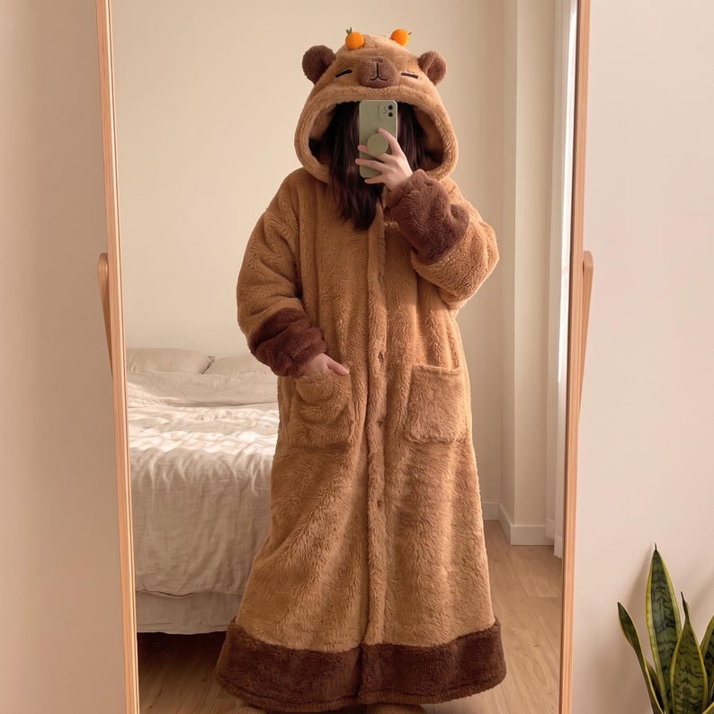 May include: A long, brown, plush fleece robe featuring a hood with embroidered capybara facial features and two small orange accents on top. The robe has long sleeves with dark brown cuffs, a button-front closure, and two large front patch pockets.