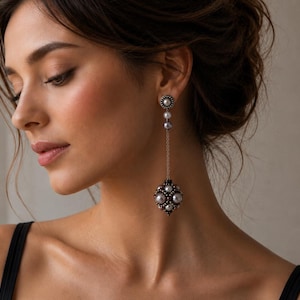 May include: A long drop earring featuring a small circular stud with a central pearl, connected by a thin chain to a larger, spherical pendant adorned with multiple grey pearls and dark metallic beads, creating a layered and intricate design.