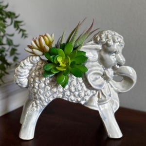 May include: A glossy, light gray ceramic planter shaped like a standing lamb with textured wool detailing. The hollow back of the lamb holds a cluster of artificial green and yellow succulent plants with pointed leaves and a small bow at the neck.