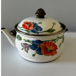 Pode incluir: Bule de esmalte creme com tampa, decorado com desenhos florais vermelhos, azuis e roxos e folhas verdes. O bule tem um bico, uma alça marrom escura e um botão na tampa.