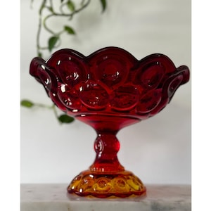 May include: A ruby red glass pedestal bowl with a scalloped rim. The bowl features a star and moon pattern. The base is a combination of red and amber glass. The bowl is a vintage piece.