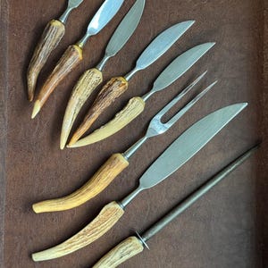 May include: A selection of vintage cutlery featuring stainless steel blades and antler handles. The set includes various knives, a carving fork, and a sharpening steel. The handles range in colour from light tan to dark brown.
