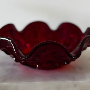 May include: A ruby red glass bowl with a ruffled edge. The bowl has a wavy, scalloped rim and a glossy finish. The bowl is likely vintage and could be used for serving or display.