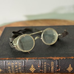 Könnte beinhalten: Antike runde Brille mit Metallrahmen und dunklen Gläsern, die auf einem alten Buch liegt. Das Buch ist in dunkles Leder gebunden und hat goldene Buchstaben und dekorative Akzente. Die Brille hat ein schwarzes Band.