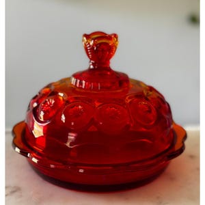 Puede incluir: Una mantequera de cristal rojo vintage con tapa. El plato tiene una base redonda y una tapa abovedada con un asa decorativa. La tapa y la base están adornadas con patrones en relieve de estrellas. El cristal tiene un acabado brillante.