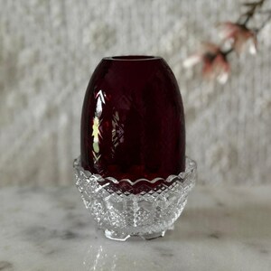 May include: A deep red glass egg-shaped object with a faceted diamond pattern resting inside a clear, colorless glass bowl base that features a scalloped rim and a textured diamond-cut design on its exterior surface.