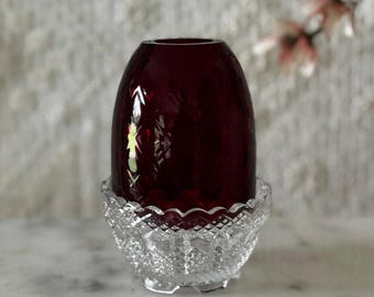 May include: A deep red glass egg-shaped object with a faceted diamond pattern resting inside a clear, colorless glass bowl base that features a scalloped rim and a textured diamond-cut design on its exterior surface.