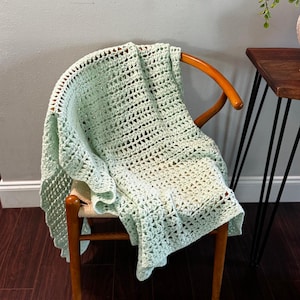 May include: A light green crocheted blanket draped over a wooden chair. The blanket has a textured, open weave design and a scalloped edge. The chair has a curved back and wooden legs.