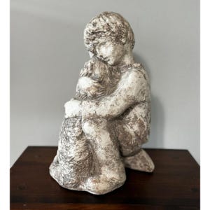 Op de afbeelding: Een gedetailleerde sculptuur van een persoon in zittende positie, die zijn knieën omhelst. De figuur is weergegeven in een neutraal kleurenpalet met getextureerde details, waardoor een verweerde uitstraling ontstaat. De sculptuur staat op een donker houten oppervlak.