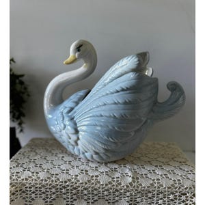 May include: A ceramic swan figurine featuring a glossy finish in shades of pale blue and white. The swan has a long, curved neck, a yellow beak, and detailed molded feathers across its body, with a hollow opening at the top of its back.