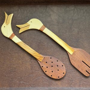 Puede incluir: Dos utensilios de cocina de madera con forma de pato. Uno es una cuchara ranurada y el otro una espátula con ranuras. Las cabezas de pato son de madera clara con detalles marrones, y los mangos son de madera más oscura.