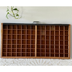Puede incluir: Una bandeja de impresora de madera vintage con numerosos compartimentos pequeños. La bandeja es rectangular con un asa oscura y un borde superior negro. El interior está dividido en una cuadrícula de pequeños espacios cuadrados y rectangulares.