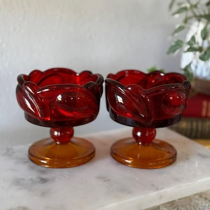 Puede incluir: Dos copas de postre de vidrio rojo vintage con un diseño decorativo de hojas alrededor del borde. Las copas tienen una base redonda de color ámbar y un pequeño tallo. El vidrio tiene un acabado brillante.