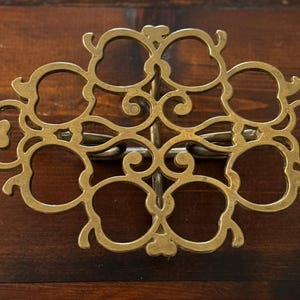 May include: Ornate brass drawer pull with a symmetrical, openwork design. The decorative hardware features a series of interconnected, stylized floral or foliate shapes. The brass finish contrasts with the dark wood surface.