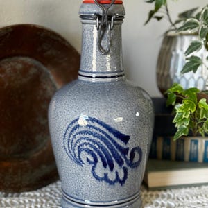 May include: A grey ceramic bottle with a white stopper and a metal clasp. The bottle has a blue and white design and is decorated with a blue wave pattern. The bottle is a decorative item.