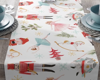 May include: A white table runner with a festive Christmas design featuring nutcracker soldiers, ballerinas, and Christmas trees. The runner is decorated with red, green, gold, and blue accents.