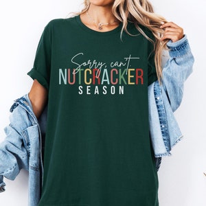 May include: Dark green t-shirt with the text "Sorry, can't. NUTCRACKER SEASON" in white, red, green, and yellow. The shirt has short sleeves and a relaxed fit. The shirt is a comfortable, casual style.