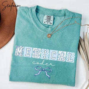 May include: Seafoam green t-shirt with the words "MEDICAL coder" in white and blue floral print. A gold necklace with a pendant rests on the shirt. The shirt also features a blue bow graphic.