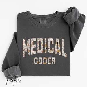 May include: Dark gray sweatshirt with the words "MEDICAL CODER" in a floral pattern across the front. The sweatshirt has long sleeves and a crew neck. The cuffs are folded up.