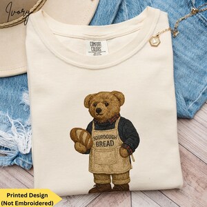 May include: Ivory-colored Comfort Colors t-shirt featuring a bear wearing an apron that says "SOURDOUGH BREAD" and holding a loaf of bread. The bear is wearing a black sweater and a red plaid shirt. The shirt is on a denim surface.
