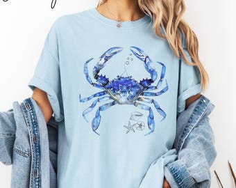 Könnte beinhalten: Ein hellblaues T-Shirt mit einer Aquarell-Illustration eines blauen Krebses, eines Seesterns und einer Muschel. Der Krebs befindet sich in der Mitte des Shirts und ist von Blasen umgeben.